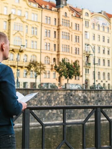 A backpacker with a blue and beige backpack stands on a bridge, admiring the row of colorful historic buildings across the river. Holding a map, they seem like a tourist exploring Europe, perhaps pondering over travel tips for their next adventure.