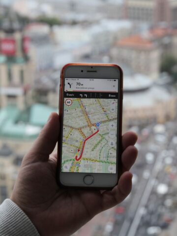 A person's hand holds a smartphone displaying a map with a marked route, perfect for the travel pro. The background reveals an aerial cityscape, bustling with buildings and roads, offering navigating tips to master foreign public transportation like a local.