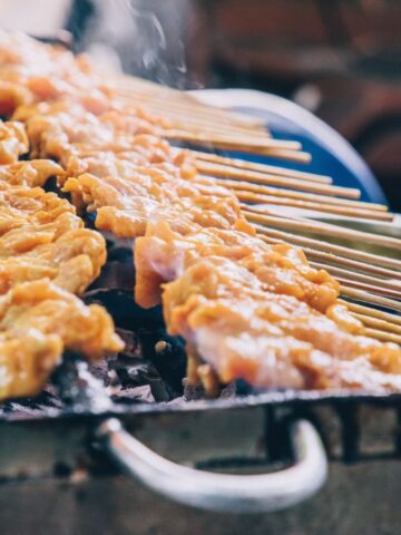 Skewers of marinated meat sizzle over an open flame, reminiscent of street foods enjoyed around the world. A hand expertly adjusts one of the skewers as fragrant smoke rises from the grill.