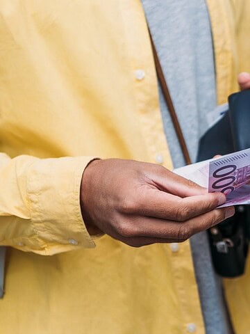 A person in a yellow jacket holds euro banknotes and a black wallet on steps, embodying the essence of traveling on a budget. The image highlights their hands and currency, underscoring savvy financial transactions or spending.