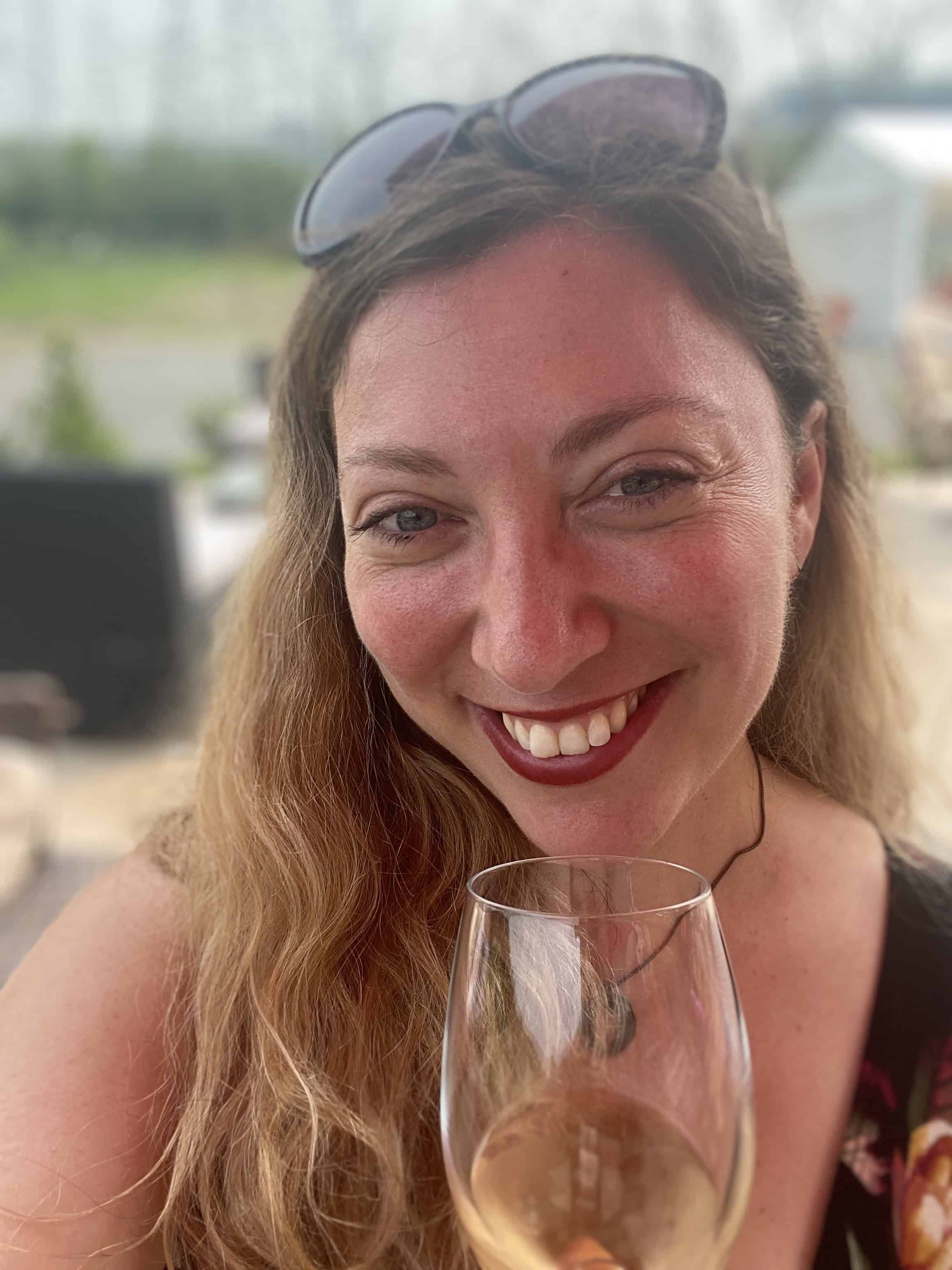 A person with long hair smiles while holding a glass of wine. Sunglasses rest on their head, capturing the joy of exploring things to do in Niagara Falls. They stand outdoors with a blurred backdrop of lush greenery and a charming building.