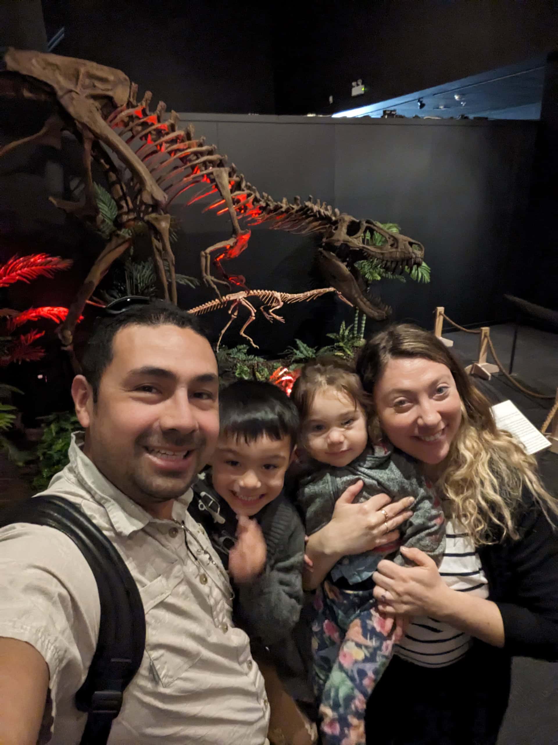 A family of four is taking a selfie in front of a dinosaur skeleton exhibit—one of the fun things to do with kids in Las Vegas. The father holds the camera while the mother holds their daughter. The son stands between them, all smiling against a dimly lit background with red accent lights.