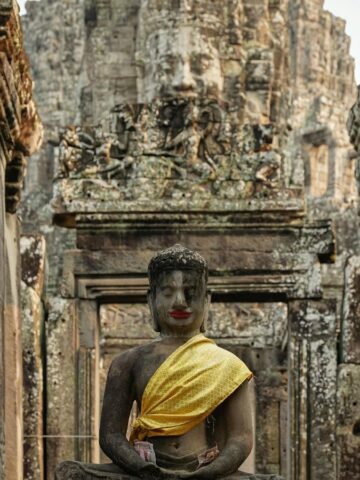 A stone statue of a seated figure with a golden sash graces the entrance of an ancient temple, offering a glimpse into the romantic allure of Southeast Asia. The temple's intricate carvings and large stone face evoke a sense of weathered history, perfect for enchanting getaways.