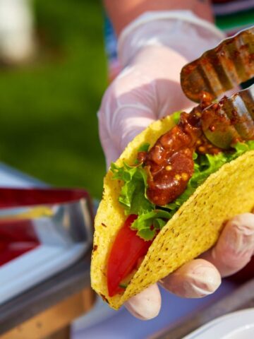 A close-up of a person holding an open hard-shell taco, a beloved street food. It's filled with lettuce, sliced tomatoes, and saucy meat. The person uses gloves and tongs to add more filling. A colorful cloth and food trays are visible in the background, evoking flavors from around the world.