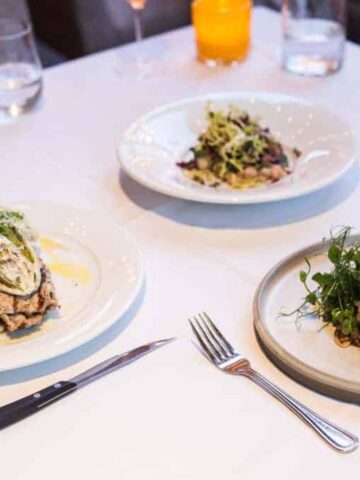 Three elegantly plated dishes are arranged on a white tablecloth at Ristorante Beatrice in Montreal. The plates include a leafy salad, a dish with what appears to be meat and sauce, and a block of food garnished with greens and placed on lentils. Utensils are set beside each plate.
