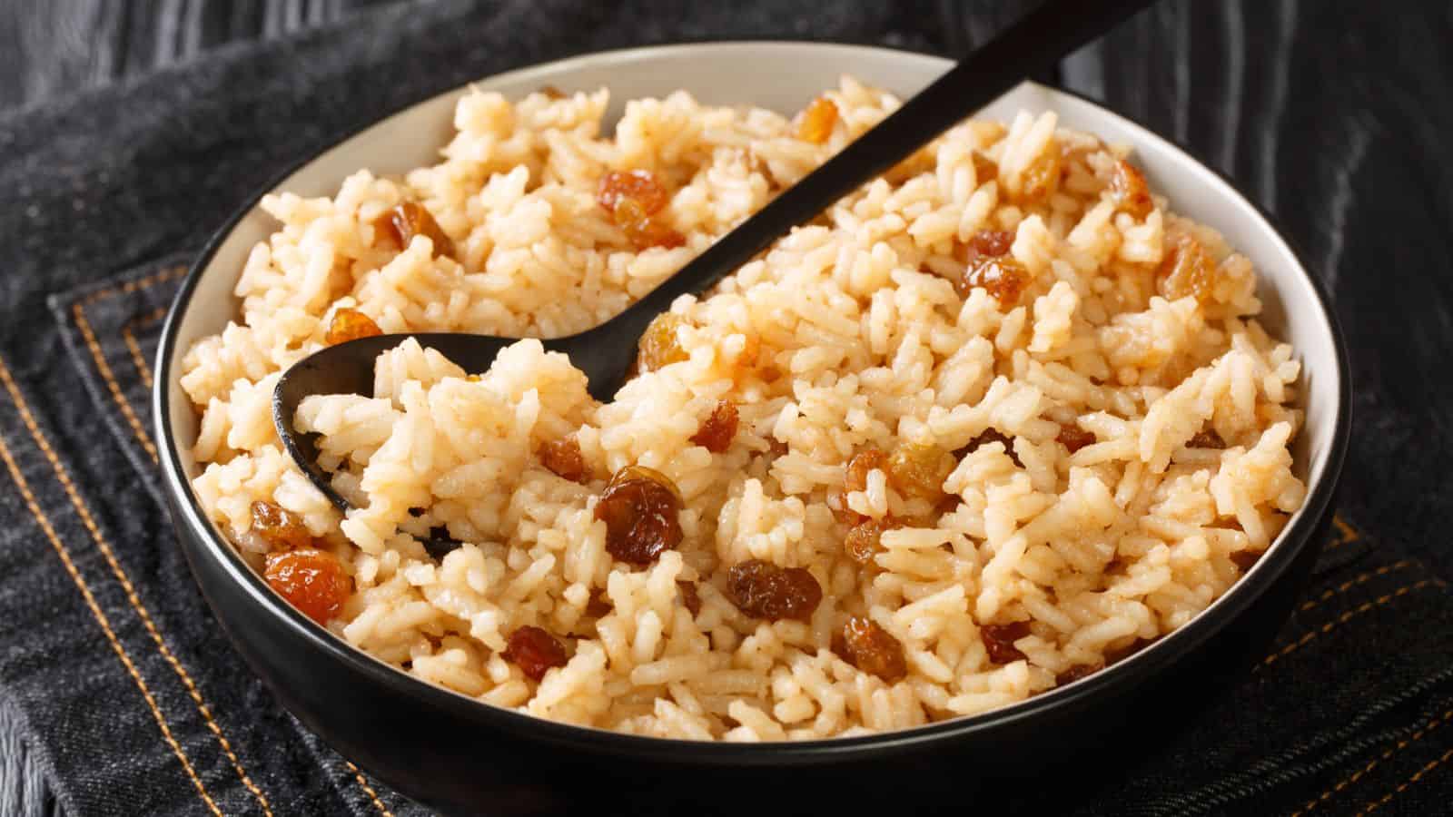 A bowl of rice pilaf with raisins is shown. The rice appears fluffy and is mixed evenly with brown raisins. A black serving spoon rests in the bowl. The bowl is placed on a dark fabric surface.