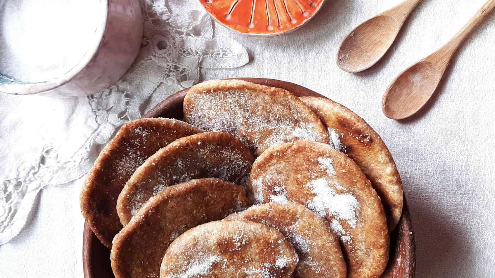 A bowl of sugar-dusted pancakes on a white lace tablecloth, accompanied by a small pink dish, two wooden spoons, an orange decorative plate, and a small cup filled with sugar.