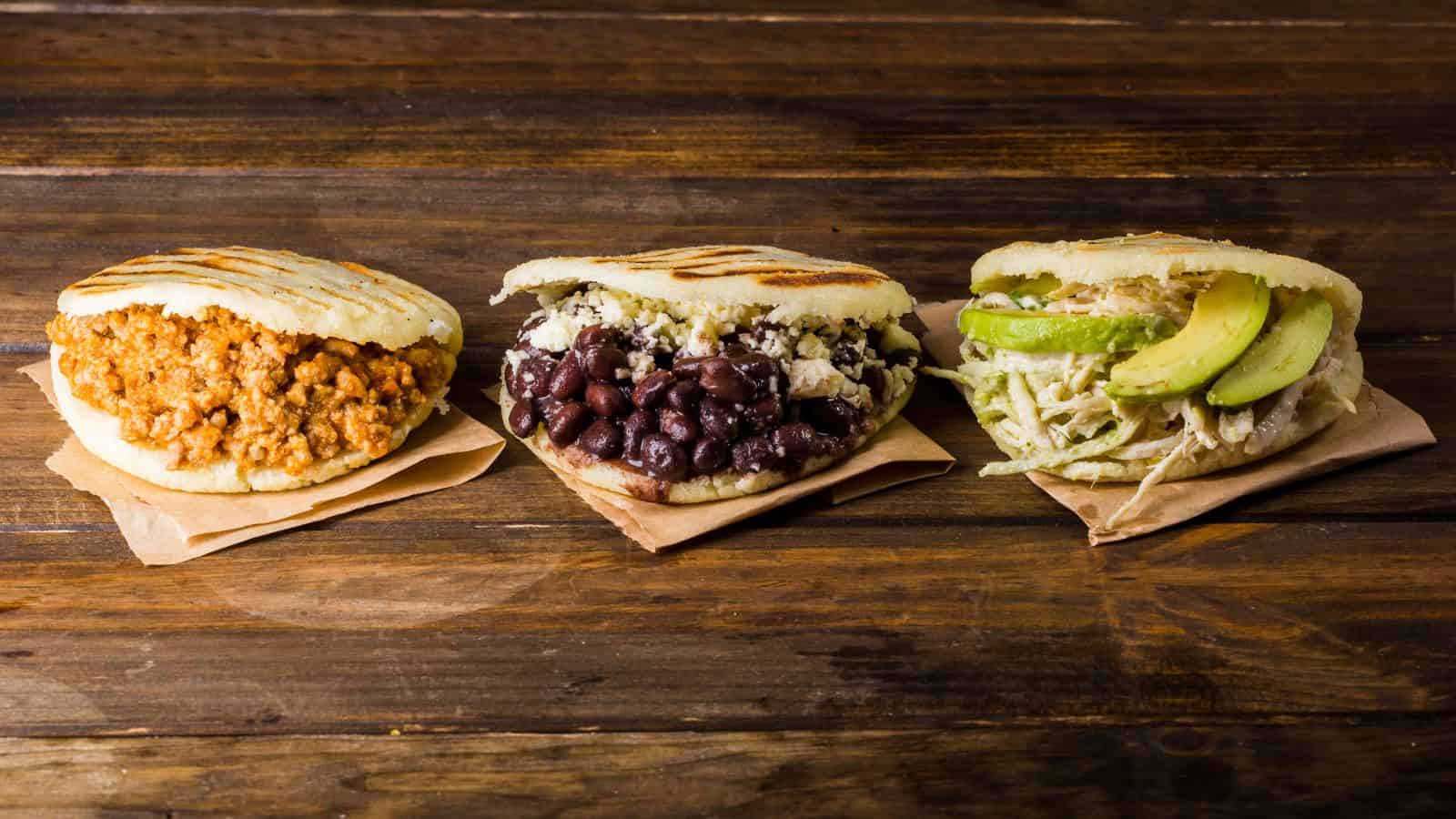 Three arepas placed on a wooden surface, each on a piece of brown paper. From left to right, they are filled with cooked minced meat, black beans and cheese, and shredded chicken with avocado slices. The arepas are slightly grilled.