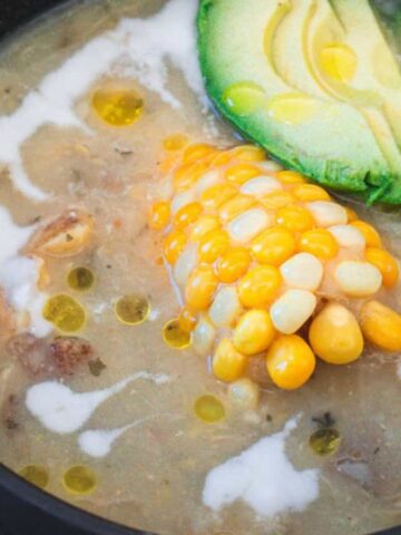 A bowl of creamy soup is topped with chunks of avocado and a piece of corn on the cob. The soup is garnished with a drizzle of white cream and olive oil, served in a dark bowl on a wooden surface. A spoon sits beside the bowl.
