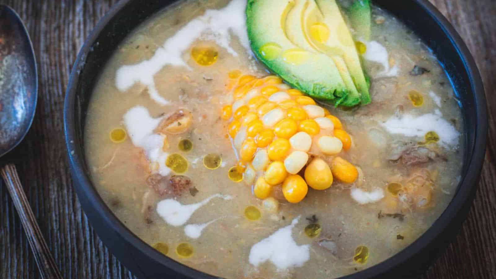 A bowl of creamy soup is topped with chunks of avocado and a piece of corn on the cob. The soup is garnished with a drizzle of white cream and olive oil, served in a dark bowl on a wooden surface. A spoon sits beside the bowl.
