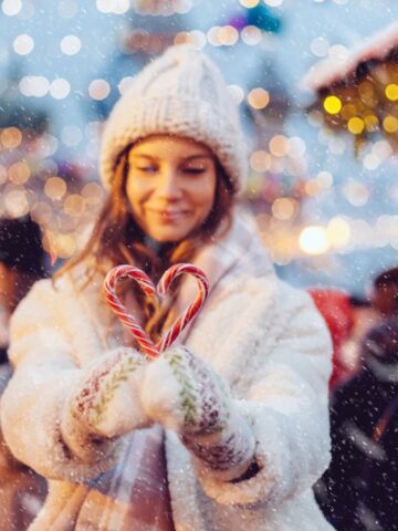 A woman in a cozy coat and knitted hat holds two candy canes shaped like a heart in her gloved hands. She stands amid winter holiday activities at a festive outdoor market, with blurred crowds and twinkling lights in the background as snow gently falls.