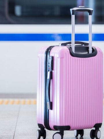 Pink hard-shell suitcase with a telescopic handle stands on a train station platform. The background features a partially visible train with a blue stripe. The suitcase has four wheels and is positioned upright.