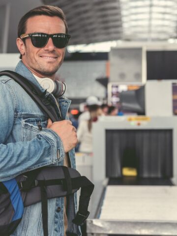 A man wearing sunglasses and a denim jacket stands at an airport security checkpoint with a blue backpack, headphones around his neck. The background shows people and security equipment.