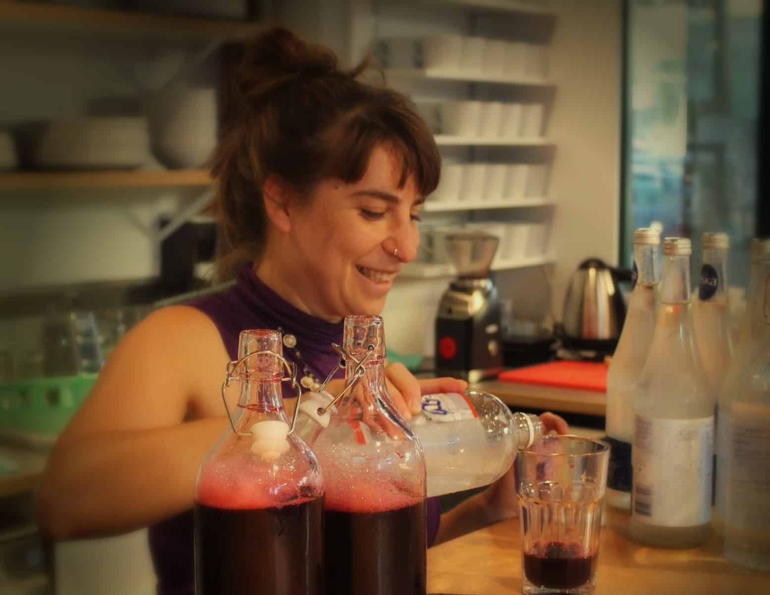 Woman smiling while pouring clear liquid into a glass with red drink, bottles and kitchenware in background—capturing the spirit of the Fletcher Beyond the Bagel Tour.