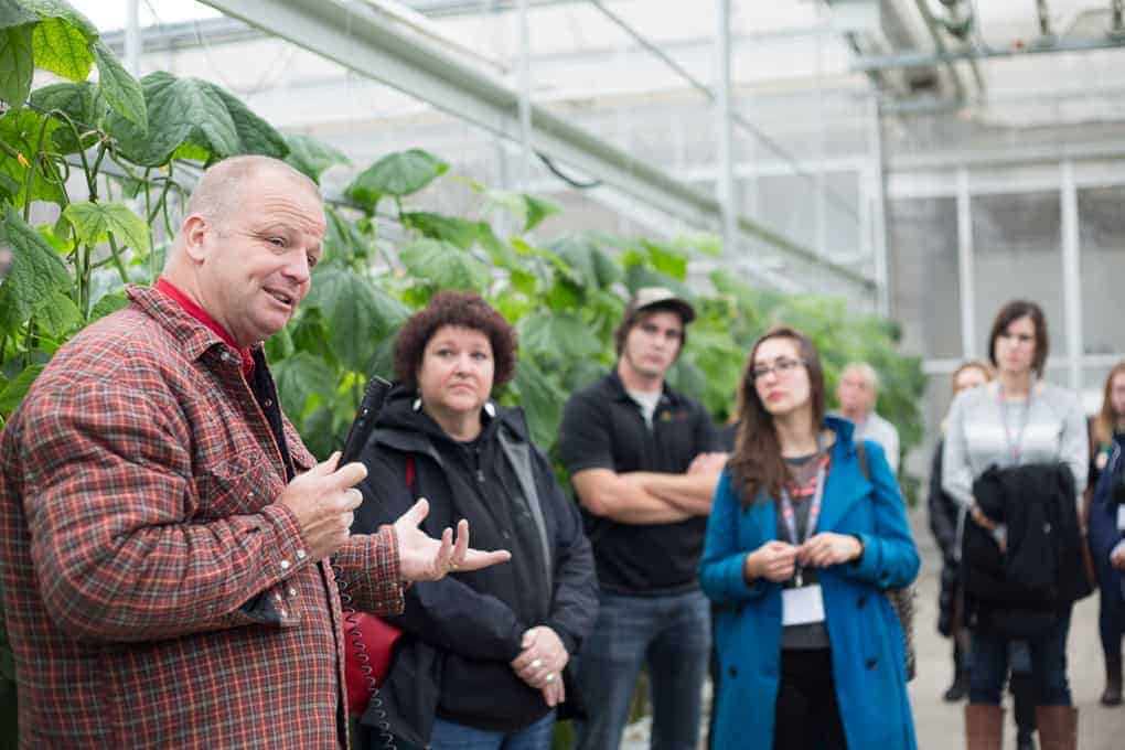A man speaks to a group of people in a greenhouse with green plants in the background, sharing insights about sustainable practices from Farm and Food Care Ontario.