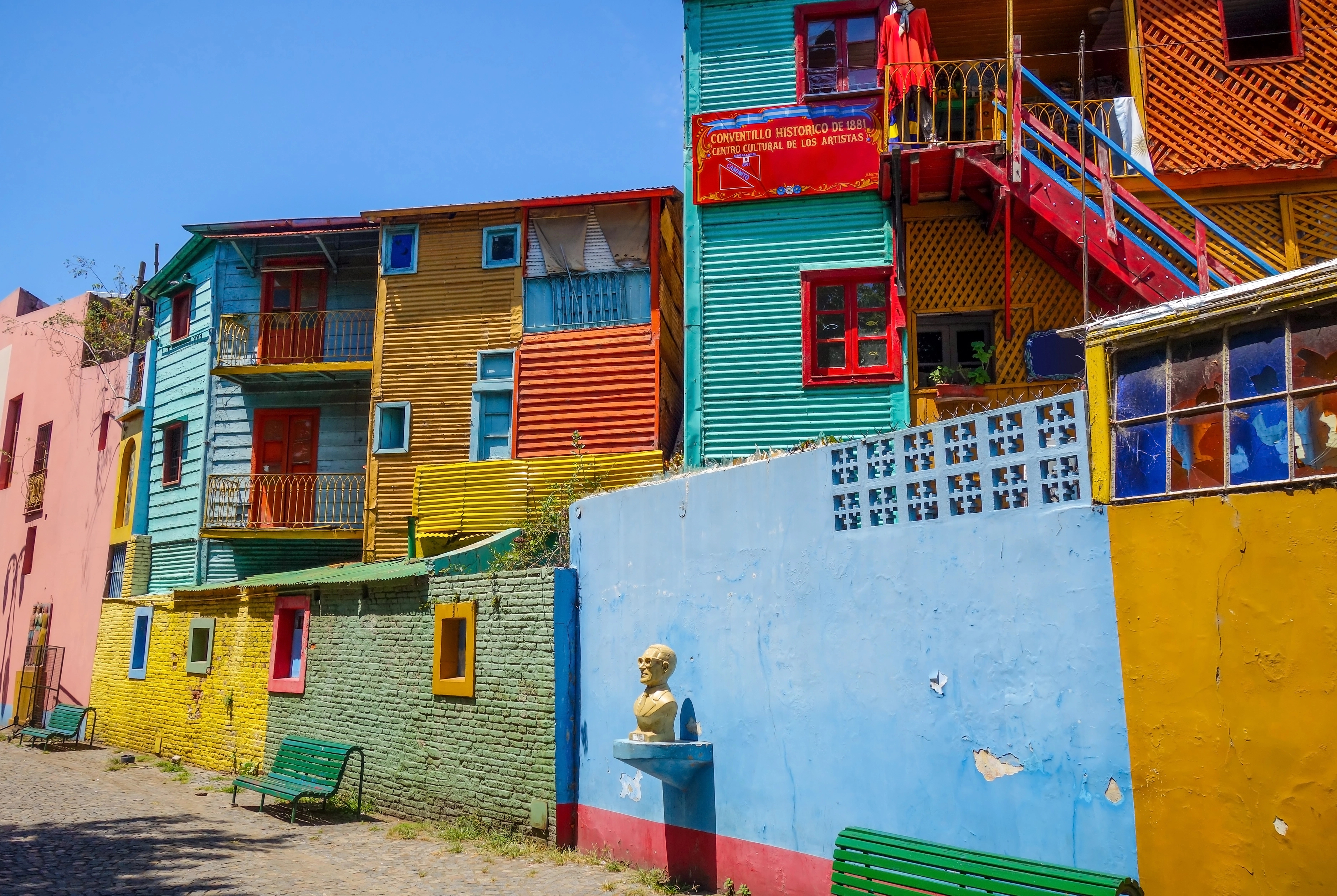 Colorful buildings with red, yellow, and blue facades line a lively street scene—complete with benches and a bust statue—offering classic sights among the top things to do in Buenos Aires.