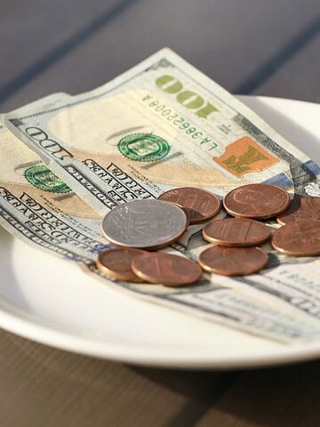 A white plate on a wooden table holds several U.S. hundred-dollar bills and various coins, including pennies and a quarter. The money is arranged casually on the plate, suggesting it might be a tip or small collection.