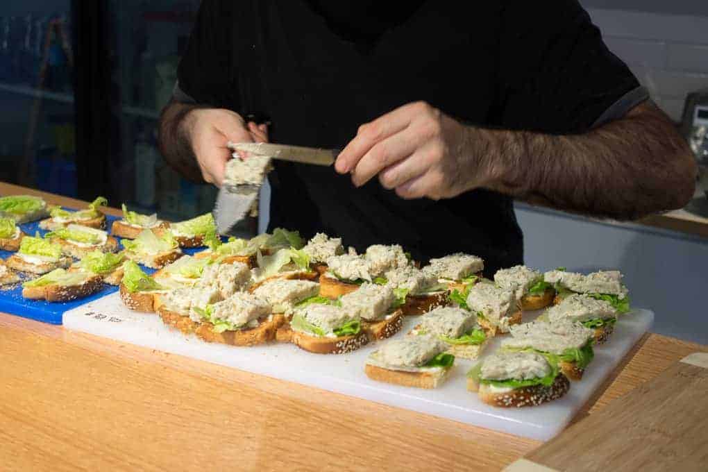 During the Fletcher Beyond the Bagel Tour, a person spreads tuna salad onto slices of bread with lettuce, preparing multiple sandwiches on a counter.