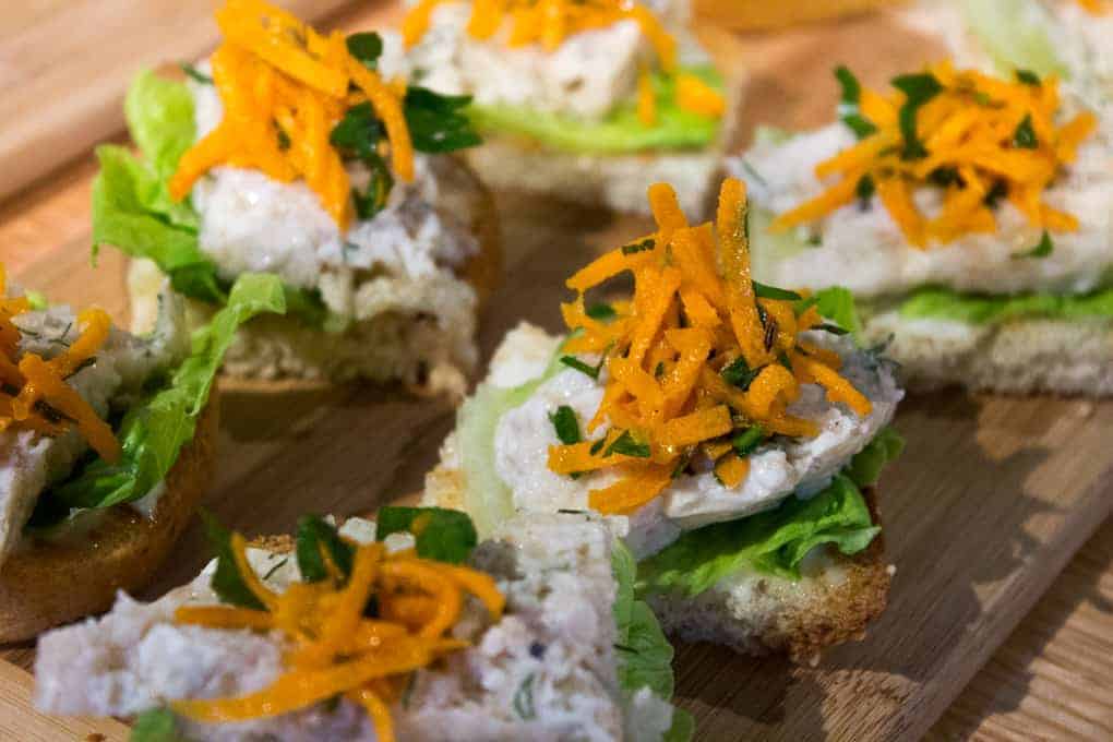 Small pieces of bread topped with lettuce, a creamy spread, and shredded carrots on a wooden board—perfect bites to enjoy during your Fletcher Beyond the Bagel Tour.