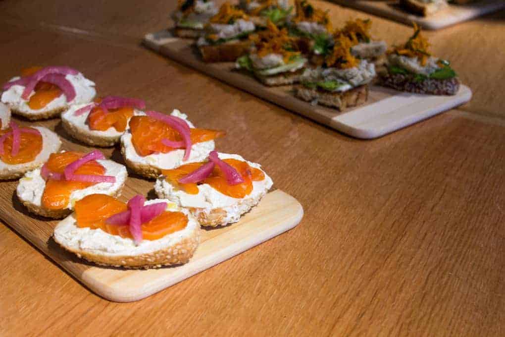 Small slices of bread with cream cheese, smoked salmon, and pickled onions on a wooden board—perfect bites inspired by the Fletcher Beyond the Bagel Tour.