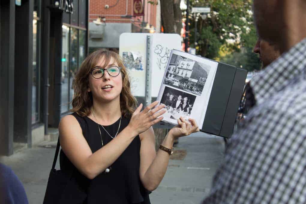 Woman with glasses leads a Fletcher Beyond the Bagel Tour, holding up photos and sharing stories as she guides a small group on an engaging street tour outdoors.