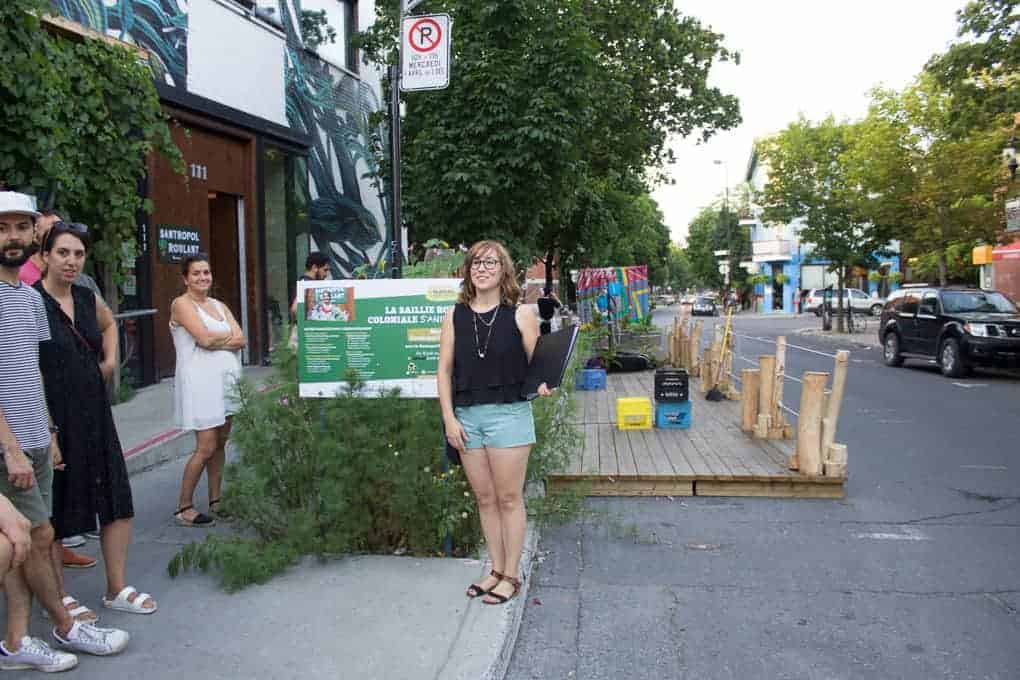 A woman stands by a green sign on a city sidewalk during the Fletcher Beyond the Bagel Tour; people and parklet structures are nearby.