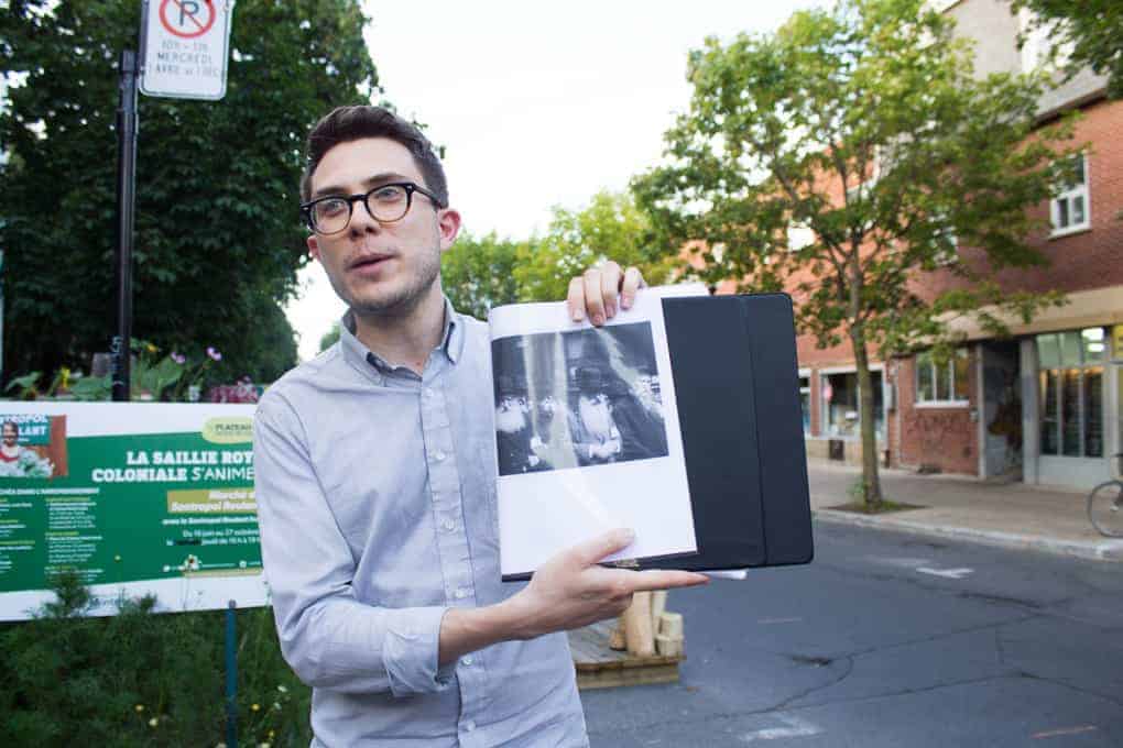 A man holds open a book with a black-and-white photo, standing on a city street near a park sign during the Fletcher Beyond the Bagel Tour.