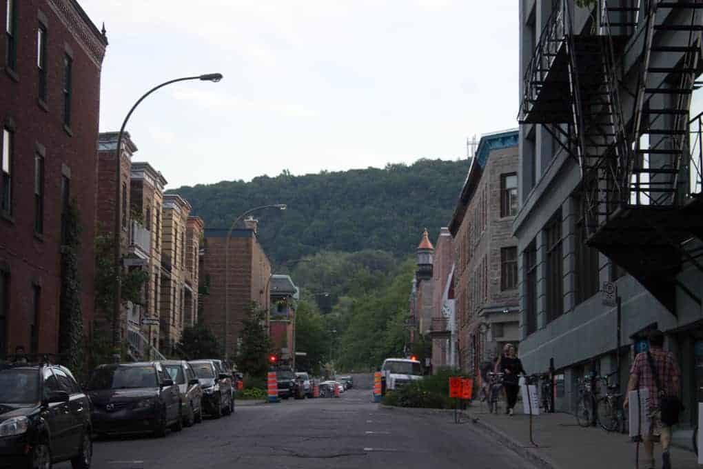 A city street with parked cars, people walking, and a green hill in the background under a cloudy sky—perfect for exploring on the Fletcher Beyond the Bagel Tour.