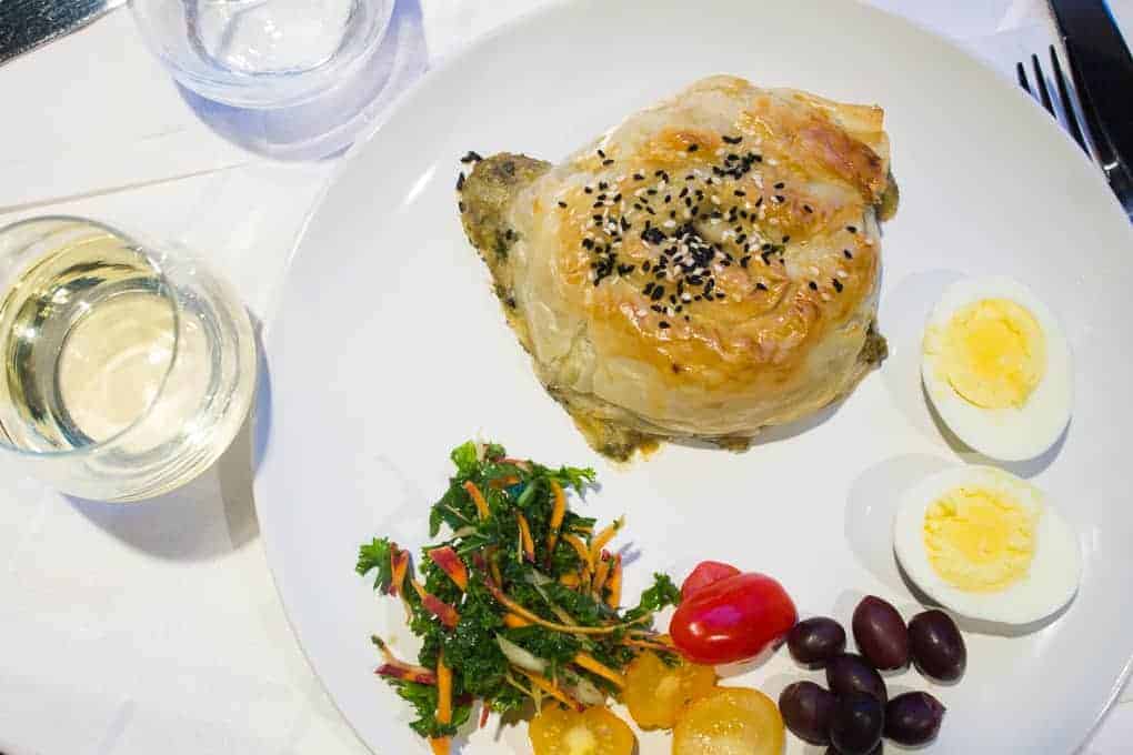 A plate with a pastry, boiled egg halves, olives, cherry tomatoes, salad, and a glass of white wine—just a taste of what you might experience on the Fletcher Beyond the Bagel Tour.