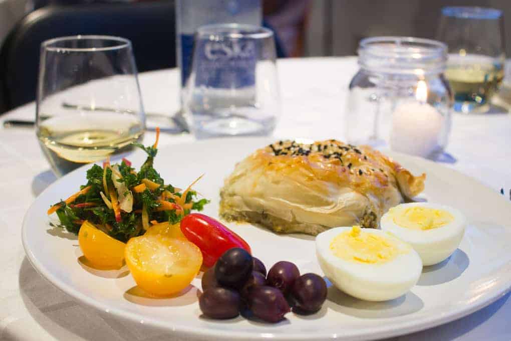 A plate with salad, boiled eggs, olives, pastry, and a glass of white wine on a table—just one of the delicious stops you might find on the Fletcher Beyond the Bagel Tour.