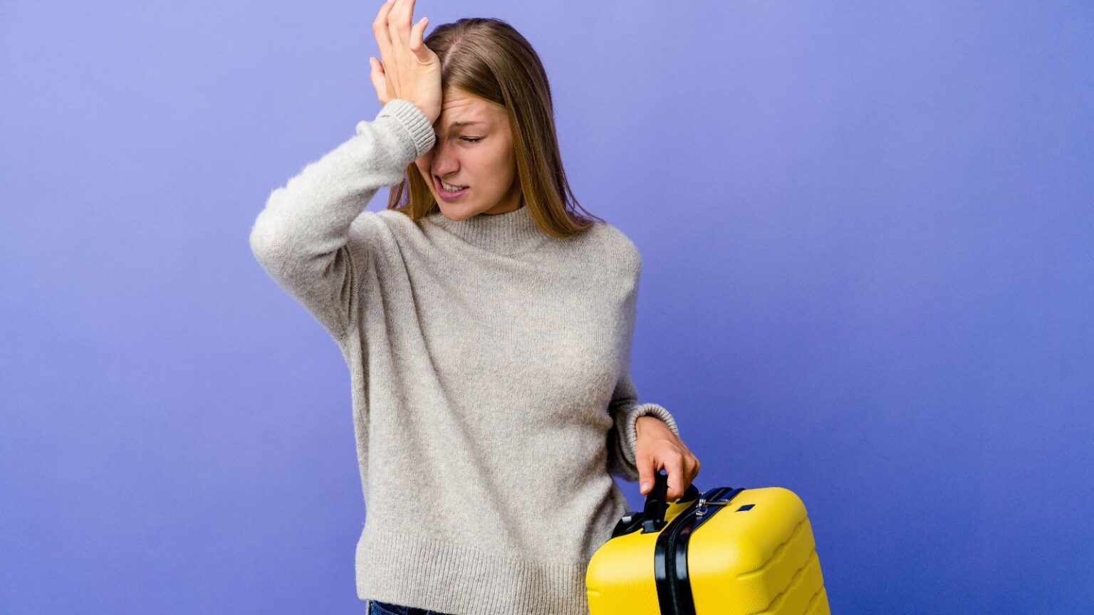 A woman in a gray sweater stands in front of a purple background, holding a yellow suitcase with one hand and touching her forehead with the other, appearing frustrated or stressed.