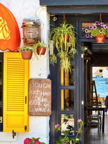 A rustic seaside restaurant with a bright orange awning and yellow shutters is adorned with potted plants and flowers. A handwritten sign suggests trying their beer and wine. Through the entry, tables are set beside a view of the blue ocean.