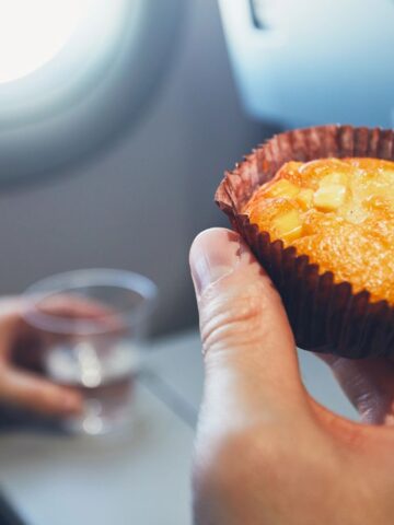 A person savors a muffin topped with cubed fruit inside an airplane, wrapped neatly in a brown paper liner. In the background, another hand clutches a clear plastic cup near the partially visible window—just a small moment in your journey to avoid common airport mistakes.