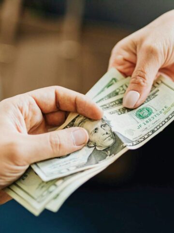 Two hands exchanging a stack of U.S. dollar bills. One person is handing over the money, and the other is receiving it. A table with a cup and scissors is partially visible in the background.