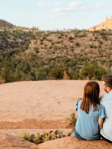 A couple sits on a large rock, overlooking a desert landscape with red rock formations and sparse vegetation under a clear sky. Both are wearing blue shirts.