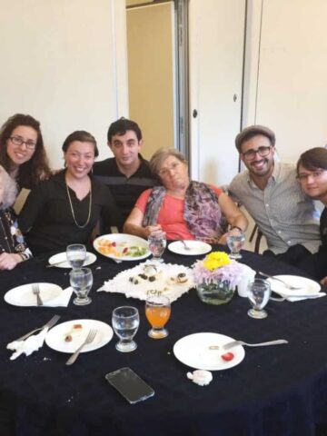 A group of seven young Jewish Argentinians is seated around a circular table with a dark tablecloth. The table has plates with food remnants, glasses, and a vase with flowers. A smartphone rests nearby. The people are smiling, posing cheerfully for the photo, crafting memories to guide their future gatherings.