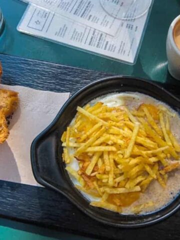 A dish of fries topped with melted cheese in a black bowl on a serving platter awaits at one of the great restaurants in Barcelona. To the side, slices of toasted bread and a cup of espresso complement the meal. A menu and water glass are visible in the background, hinting at more delights.