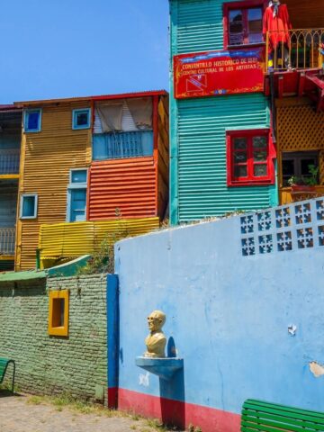 Colorful, wooden buildings with balconies and a bust statue on a blue wall line a vibrant cobblestone street—an iconic sight recommended by the best Argentina travel guide.