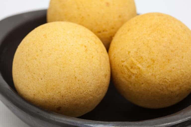 Three round, golden-brown cornmeal dumplings, reminiscent of Colombian street food, are arranged in a black bowl. The dumplings have a smooth texture and are evenly shaped, with minimal surface imperfections. The bowl is set against a plain white background.