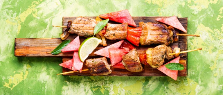 Grilled skewers with chunks of meat and vegetables, reminiscent of Colombian street food, are arranged on a wooden serving board. They are garnished with sliced watermelon, mint leaves, and a wedge of lime against a textured green background.