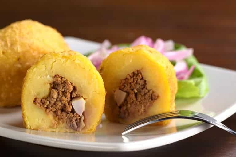 A white plate showcases a cut potato ball stuffed with ground meat and small egg pieces, embodying a taste of Colombian street food. A fork rests nearby, while sliced red onions and lettuce accompany the dish on a dark wooden surface.