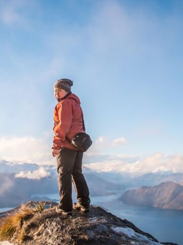 Person wearing a jacket and beanie stands on a rocky mountain peak, overlooking a scenic view of a lake and distant mountains under a clear blue sky. A camera bag hangs from their shoulder.