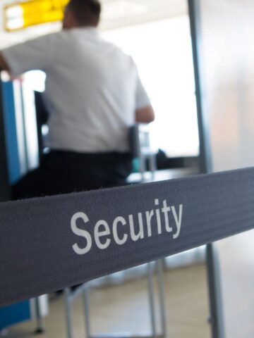 A person in a security uniform is seated at a desk. In the foreground, a fabric barrier with the word "Security" is visible, dividing the area. The setting appears to be an airport or similar transportation hub.
