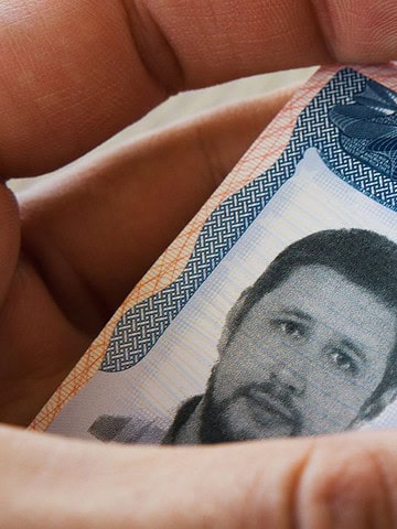 Close-up of a person holding a U.S. visa page inside a passport. The visa has a photo, name, and other personal details against a background with security patterns. The top edge shows part of the passport logo with the words "United States of America.