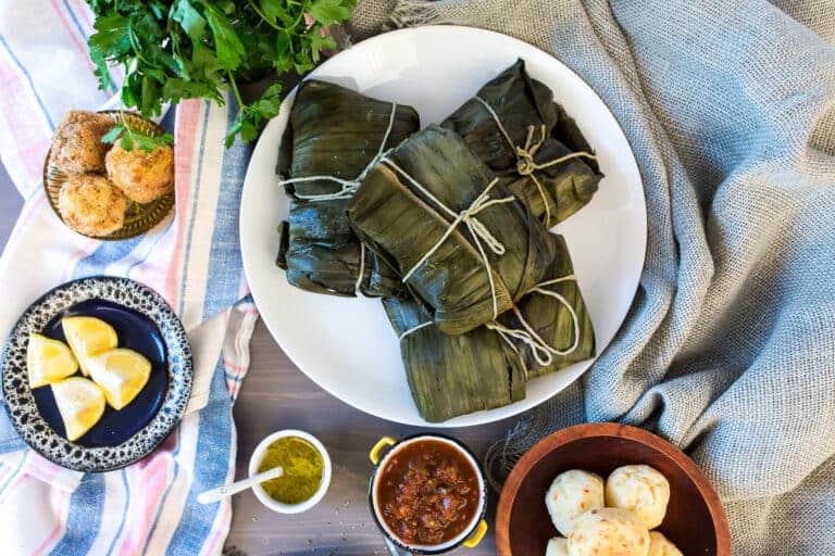 A plate of Colombian street food features tamales wrapped in green leaves and tied with string. Surrounding the plate are bowls of sauce, lemon wedges, and cheese breads. Nearby, a bunch of fresh herbs rests on a textured cloth and wooden surface.