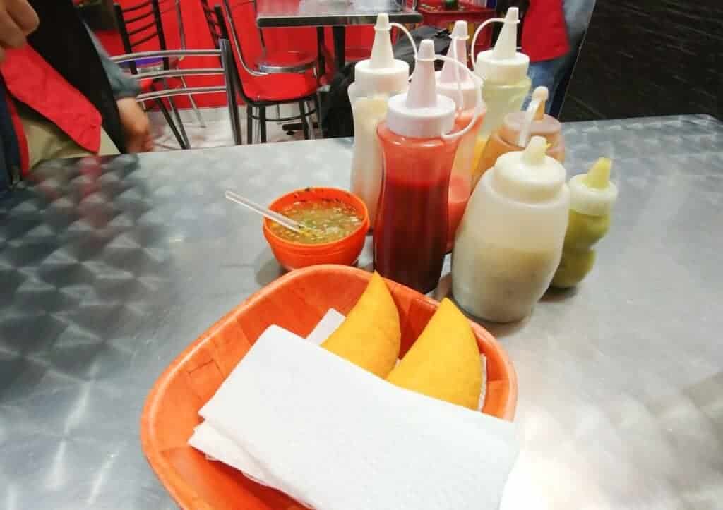 A metal table features a red dish with two empanadas on napkins, embodying authentic Colombian street food. Various condiment bottles, including ketchup and mayonnaise, sit beside a small bowl of sauce with a spoon. Red and black chairs add to the vibrant scene in the background.