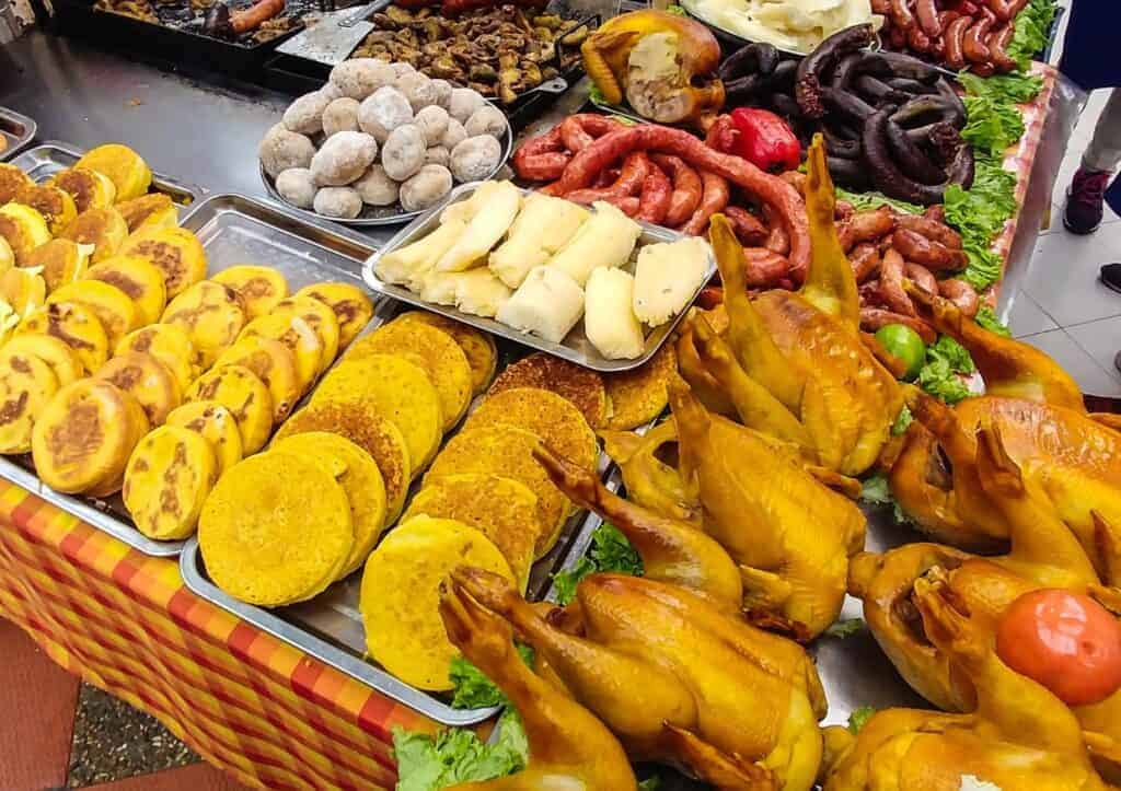 Trays of Colombian street food, including whole roasted chickens, arepas, sausages, boiled potatoes, and tamales, are displayed on a table covered with a checkered cloth.