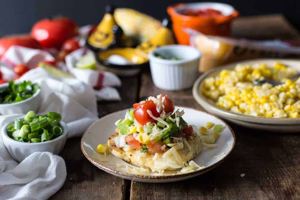 A plated dish reminiscent of Colombian street food features shredded cheese, cherry tomatoes, and herbs atop a piece of bread. Surrounding the plate are bowls of green onions, corn kernels, diced tomatoes, lime slices, and fresh tomatoes on a rustic wooden table.