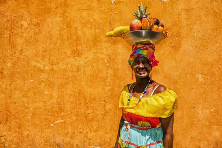 A person stands against an orange textured wall, wearing a colorful dress with a yellow top and multicolored skirt. They gracefully balance a metal bowl of exotic Colombian fruits, including bananas and pineapples, on their head and wear a multicolored headscarf.