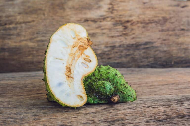 A cut soursop fruit, one of the exotic Colombian fruits, is placed on a wooden surface. The green, spiky exterior is partially visible, revealing the white, fibrous interior with embedded seeds. The background showcases a textured wooden surface.